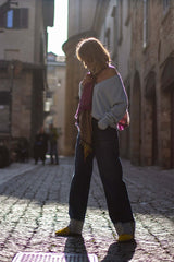 Woman wearing a loose cashmere sweater and jeans, standing on a cobblestone street, illuminated by sunlight.