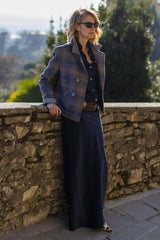 Woman in a stylish peacoat and dark skirt, posing by a stone wall in a sunny outdoor setting.