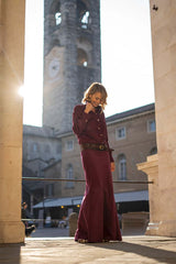 Stylish woman in a long plum velvet dress, standing against a sunlit historical building.