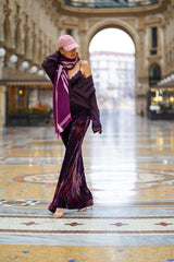 Fashion model wearing a stylish purple off-the-shoulder dress, cardigan, and pink cap in an elegant arcade.
