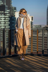 Stylish woman in a coat and wide-leg pants, wearing sunglasses and a scarf, standing on a rooftop with city skyline.