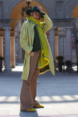 Model wearing a bright yellow boucle coat with a green scarf and wide-legged pants, set in an outdoor urban backdrop.