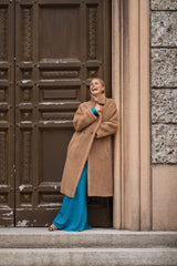 Woman in a camel teddy coat and blue dress, smiling beside an ornate door, showcasing effortless elegance.