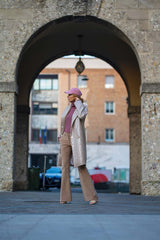 Woman in a stylish outfit wearing a beige overcoat and pink hat, walking through a city archway.