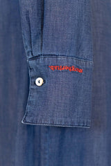 Close-up of the cuff of a denim shirt with red logo embroidery and a button detail.