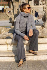 Woman in gray outfit with scarf, seated on a fountain, exuding casual elegance outdoors.
