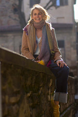 Woman wearing a cashmere scarf, smiling while sitting on a stone wall in a sunny urban setting.