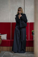 Woman in a stylish black cropped jacket and wide-leg pants, smiling against a chic red and beige backdrop.