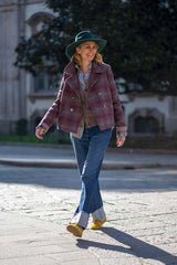 Woman in a stylish plaid jacket and green hat walking on a sunny street with a relaxed smile.