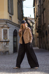 Fashionable woman in a stylish beige jacket and wide black pants walking through a cobblestone street.