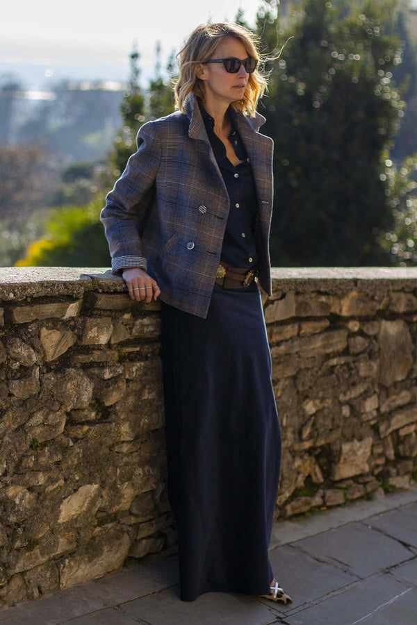 Woman in a stylish peacoat and dark skirt, posing by a stone wall in a sunny outdoor setting.