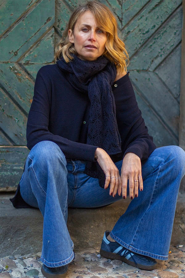 Woman seated near a green door, wearing a navy cashmere scarf and flared jeans, showcasing casual elegance.