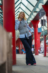 Woman smiling in a stylish cashmere gilet and striped shirt, standing confidently in a vibrant urban setting.