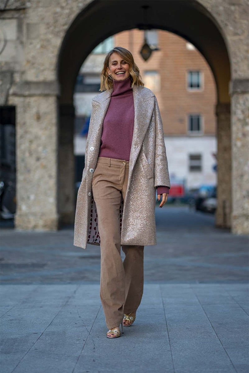 Woman in a stylish glittering coat and turtleneck sweater walking confidently through an archway.